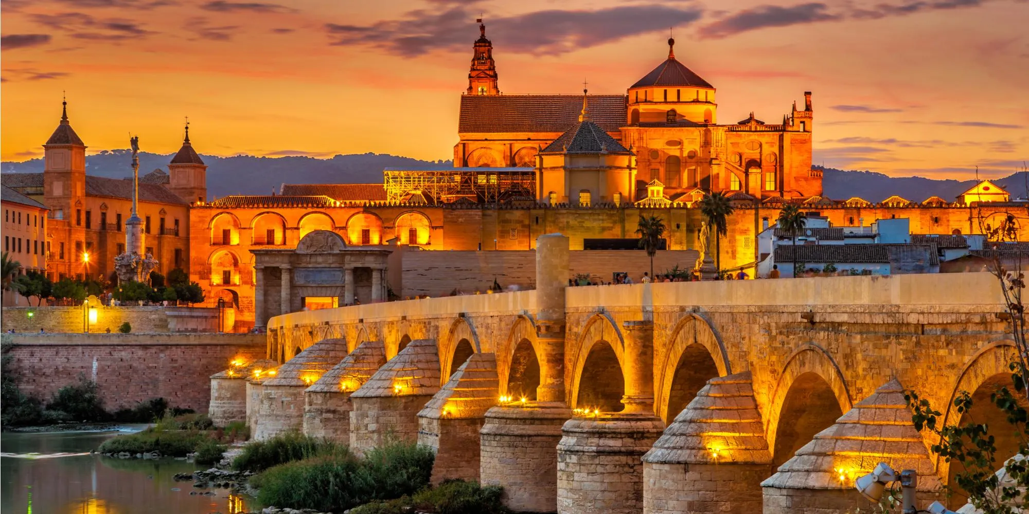 Córdoba at dusk: the Roman bridge lit gold over the Guadalquivir, and behind it the Mezquita-Catedral and its bell tower under an orange sky