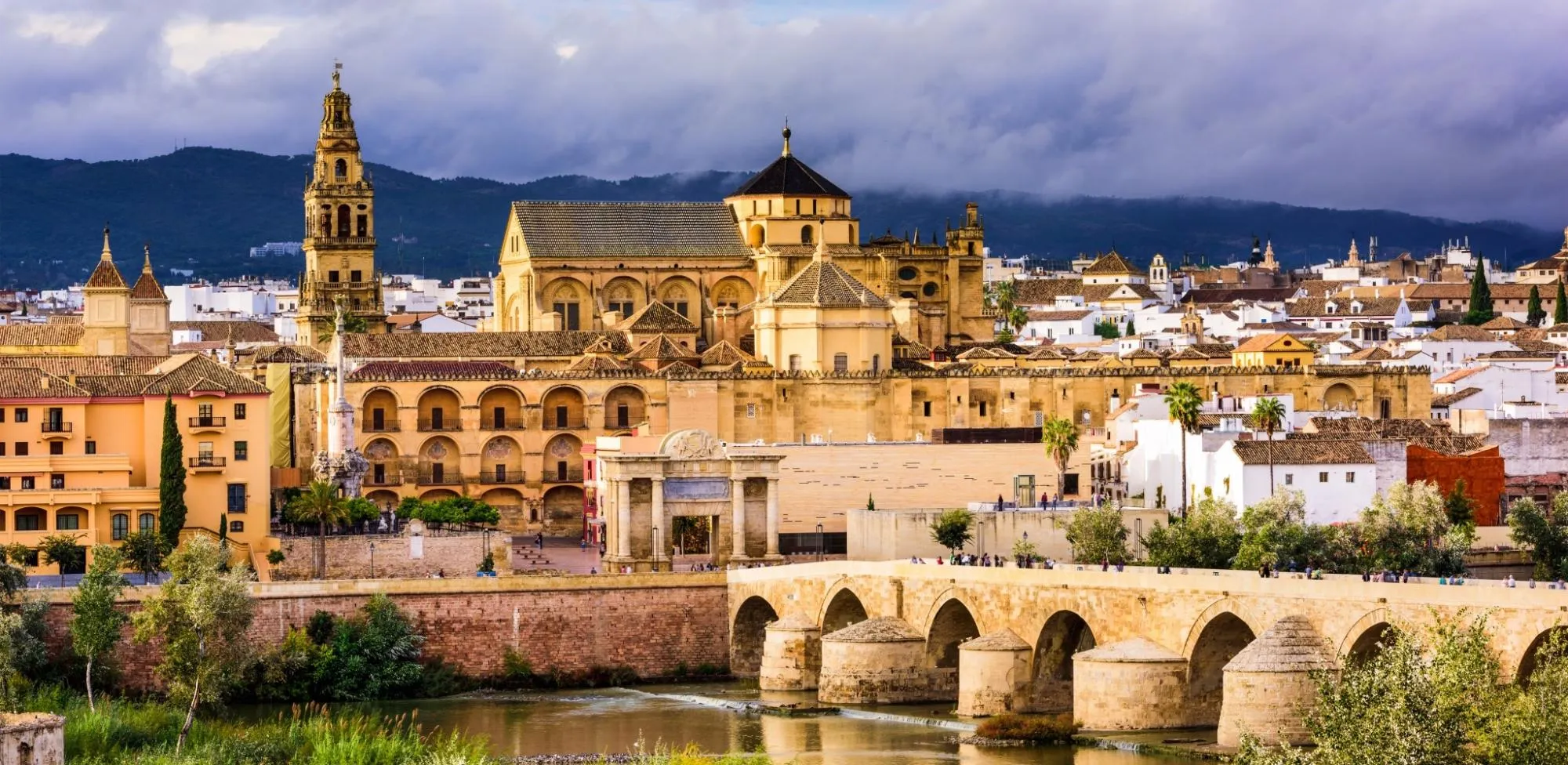 The Mezquita-Catedral above the old town of Córdoba under a stormy sky: the bell tower on the left, the cathedral's roofs and lantern rising from the long walls, a stone gateway, and the arches of the Roman bridge over the river in front, with dark hills behind