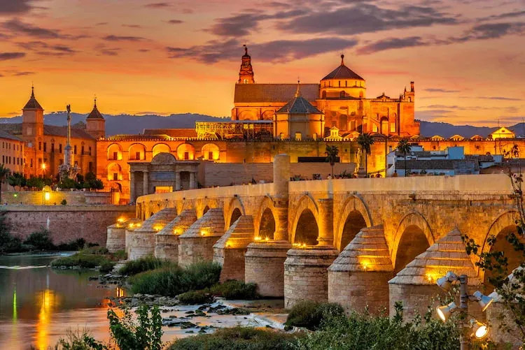 Córdoba's Roman bridge lit at dusk, with the Mezquita-Catedral rising behind it under an orange sky