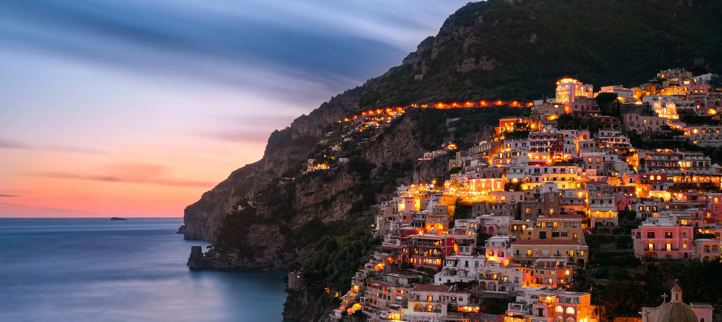 Positano at dusk, its houses lit gold and stacked up a dark, wooded cliff above a calm sea