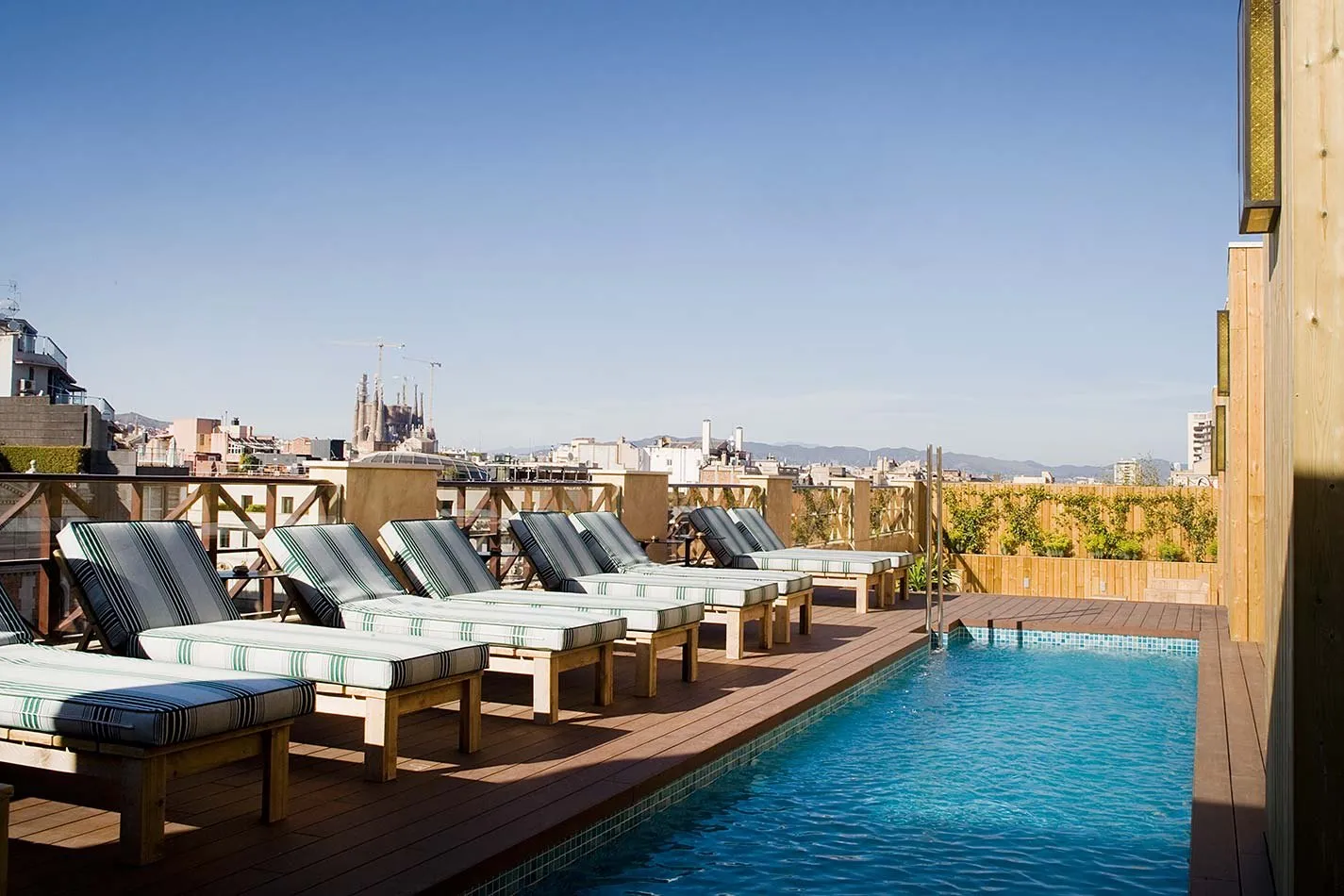 Cotton House's rooftop pool beside a timber deck lined with striped loungers, and beyond the rooftops of the Eixample the towers and cranes of the Sagrada Família, with hills on the horizon
