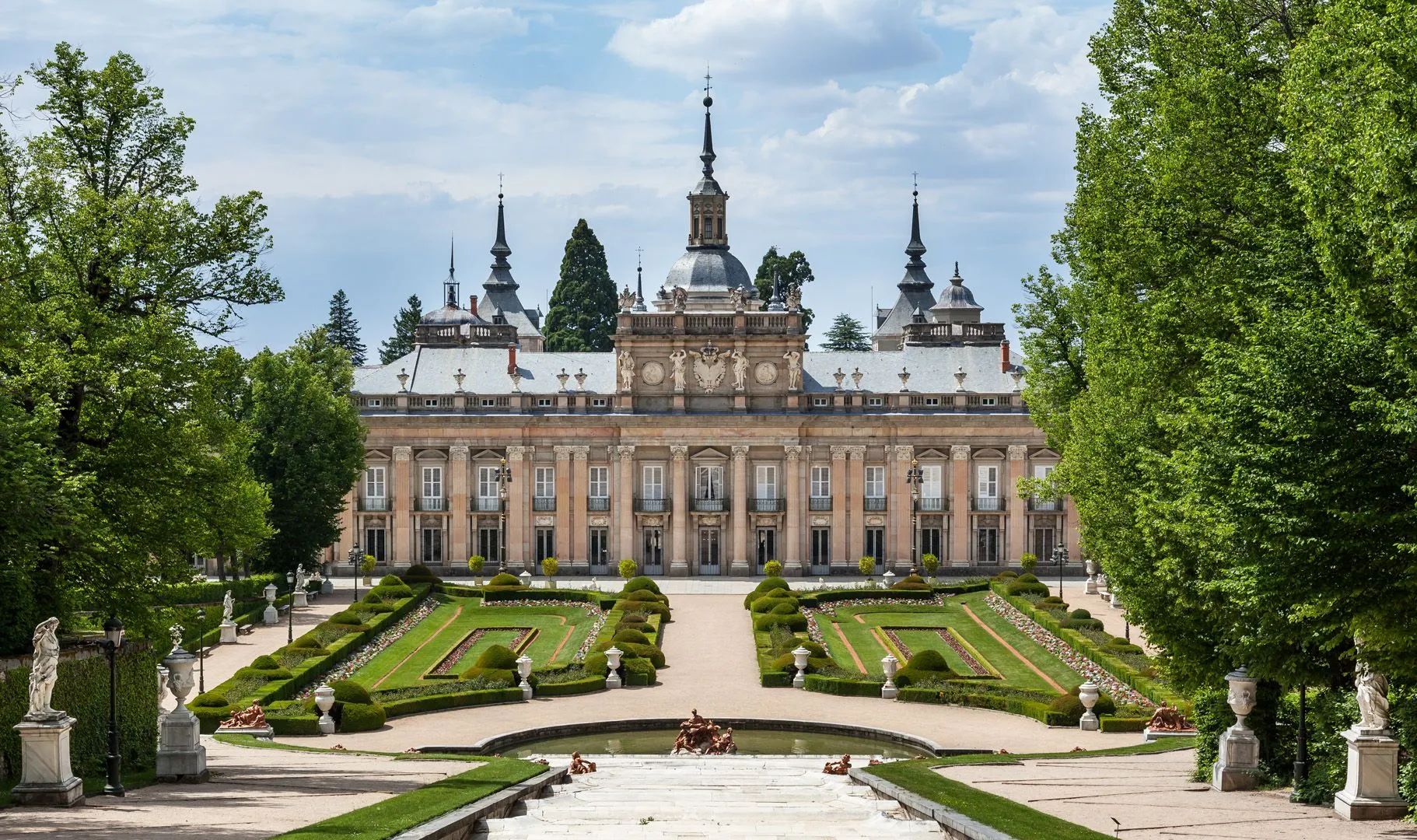The garden front of La Granja's palace, a central dome and slate spires above a long pale façade, seen across clipped parterres, a fountain basin with lead figures and white statues on pedestals between tall trees