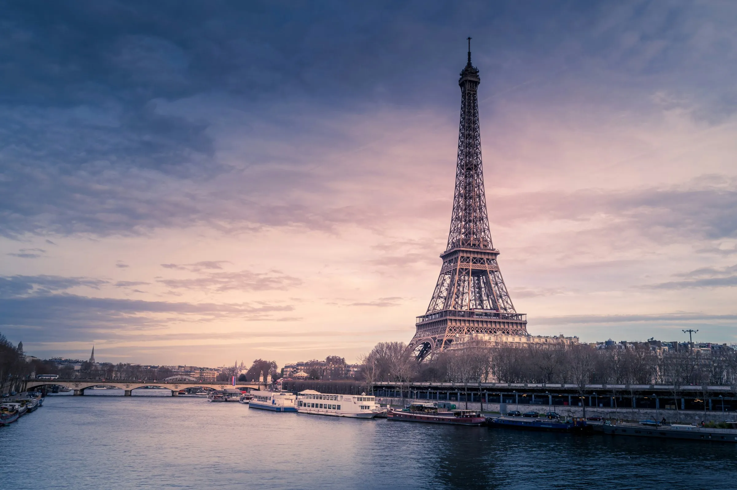 The Eiffel Tower seen across the Seine from the Right Bank at dusk, the whole tower against a mauve and pink sky, excursion boats moored along the far quay below it, the stone arches of the Pont d'Iéna to the left and bare winter trees along the bank