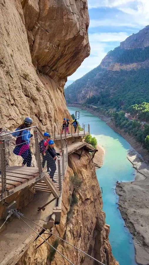 Walkers in helmets on a boardwalk pinned to the sheer orange rock wall of a gorge, a steel cable beside them and a turquoise river far below between wooded slopes