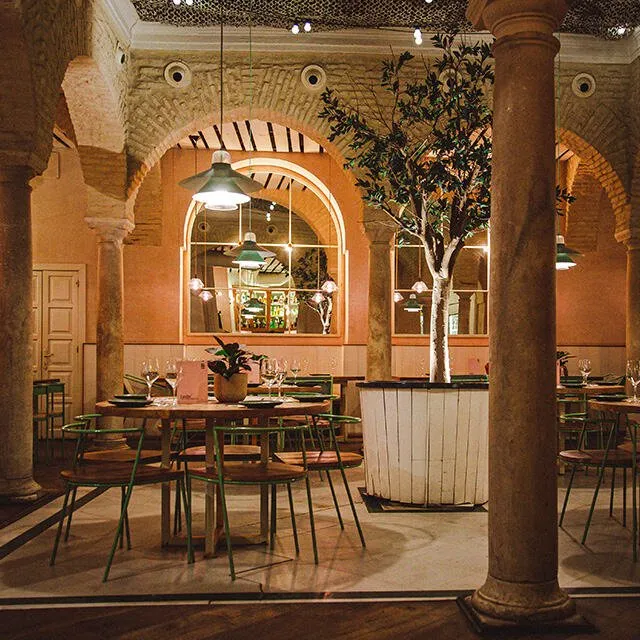 A dining room of brick arches and stone columns around a potted olive tree, with green metal chairs at round tables under hanging lamps