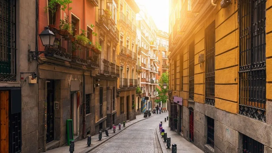 A narrow cobbled street between tall ochre and stone houses with iron balconies and potted plants, lit by a low sun at its far end