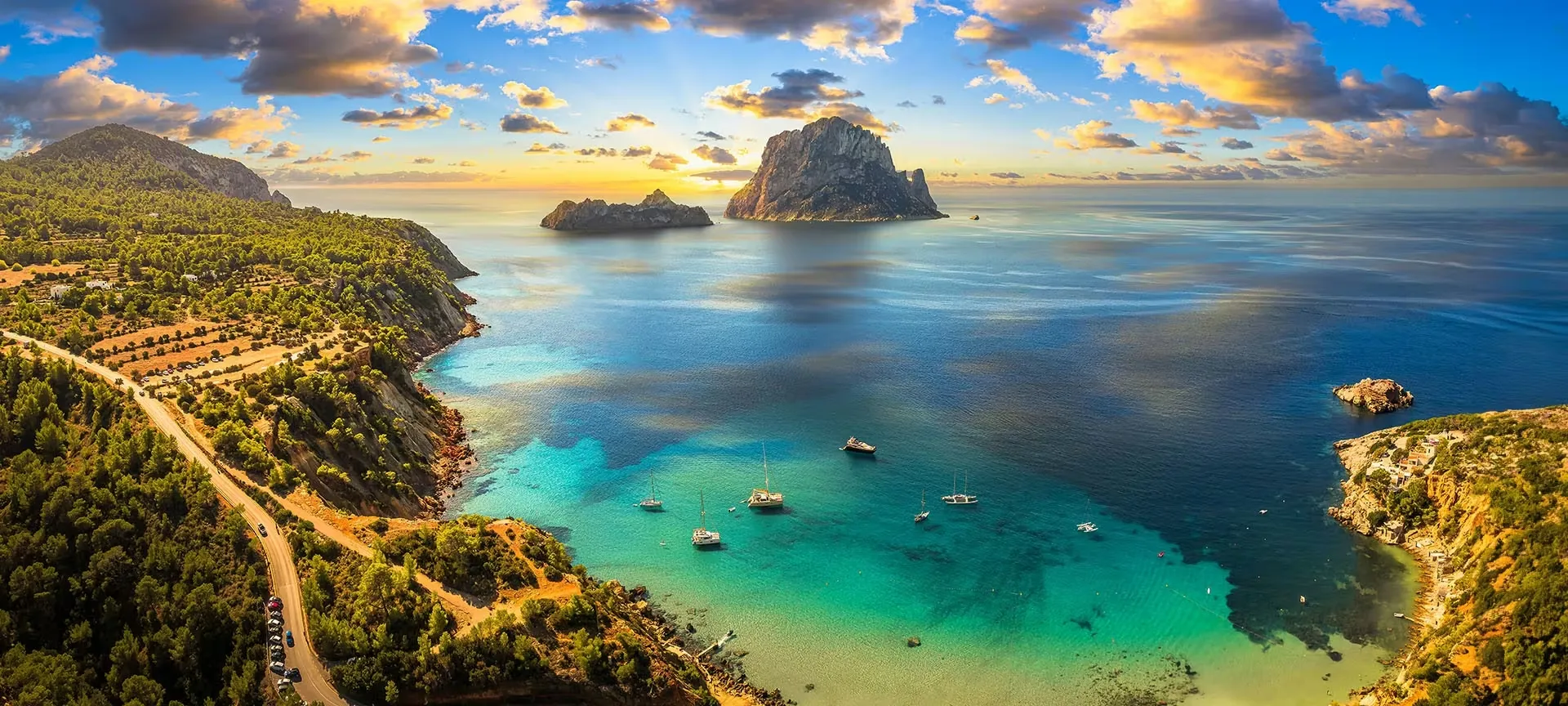 Es Vedrà and the smaller Es Vedranell at sunset, seen from high above a turquoise bay on Ibiza's south-west coast, with yachts at anchor below pine-covered cliffs and a coast road winding along them