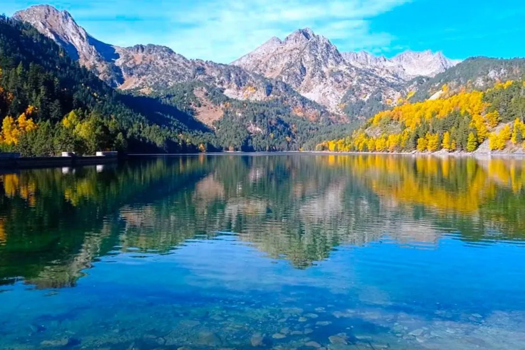A still mountain lake reflecting dark pine woods, autumn-gold trees and rocky grey peaks under a blue sky, a low dam wall at the left