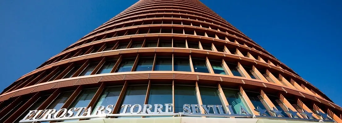 Torre Sevilla seen from its foot against a clear blue sky, the copper-coloured fins of its façade curving away overhead above the hotel's name
