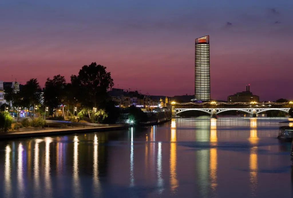 Torre Sevilla lit at dusk above the Guadalquivir, its glass shaft rising beyond the lamps of the Triana bridge, with the river carrying their reflections