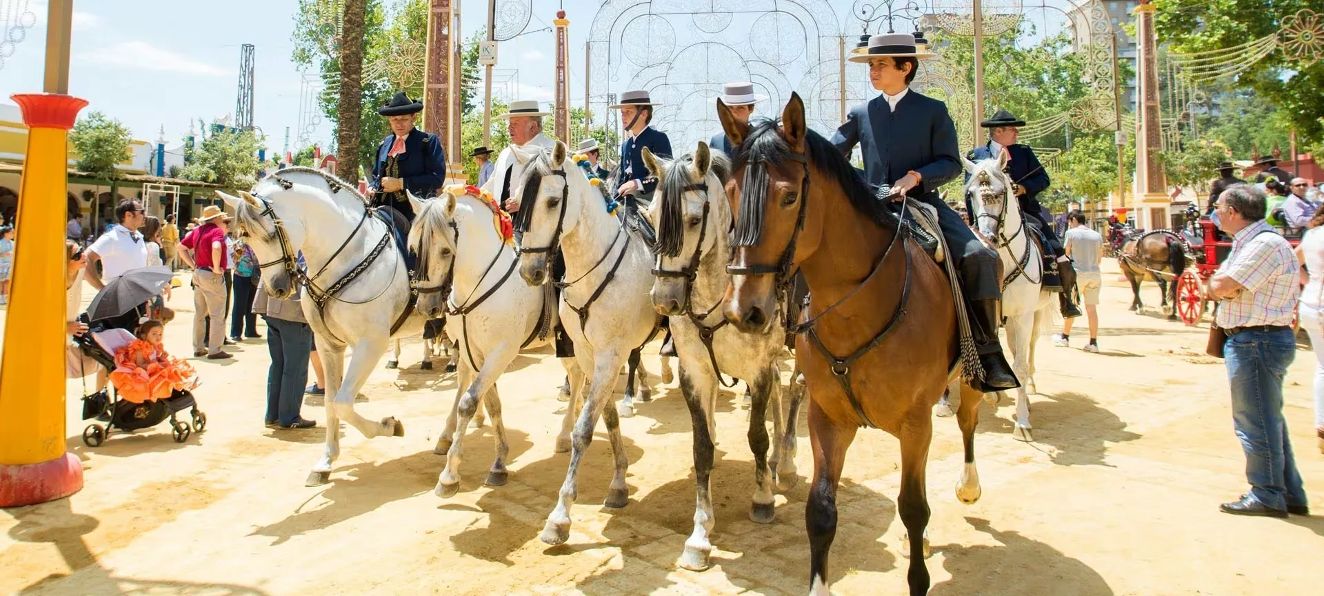 Riders in short jackets and flat-brimmed hats on grey and bay horses walking abreast down a sandy avenue of the fairground, beneath arches of festival lights, with onlookers at the sides and a small girl in an orange flamenco dress in a pushchair