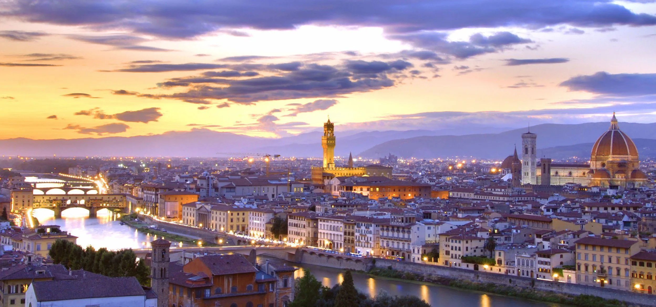 Florence at dusk under a violet and gold sky: the Arno and its lit bridges on the left, the Ponte Vecchio among them, the tower of the Palazzo Vecchio rising from the rooftops in the middle, and the Duomo's dome and Giotto's bell tower on the right, with hills beyond