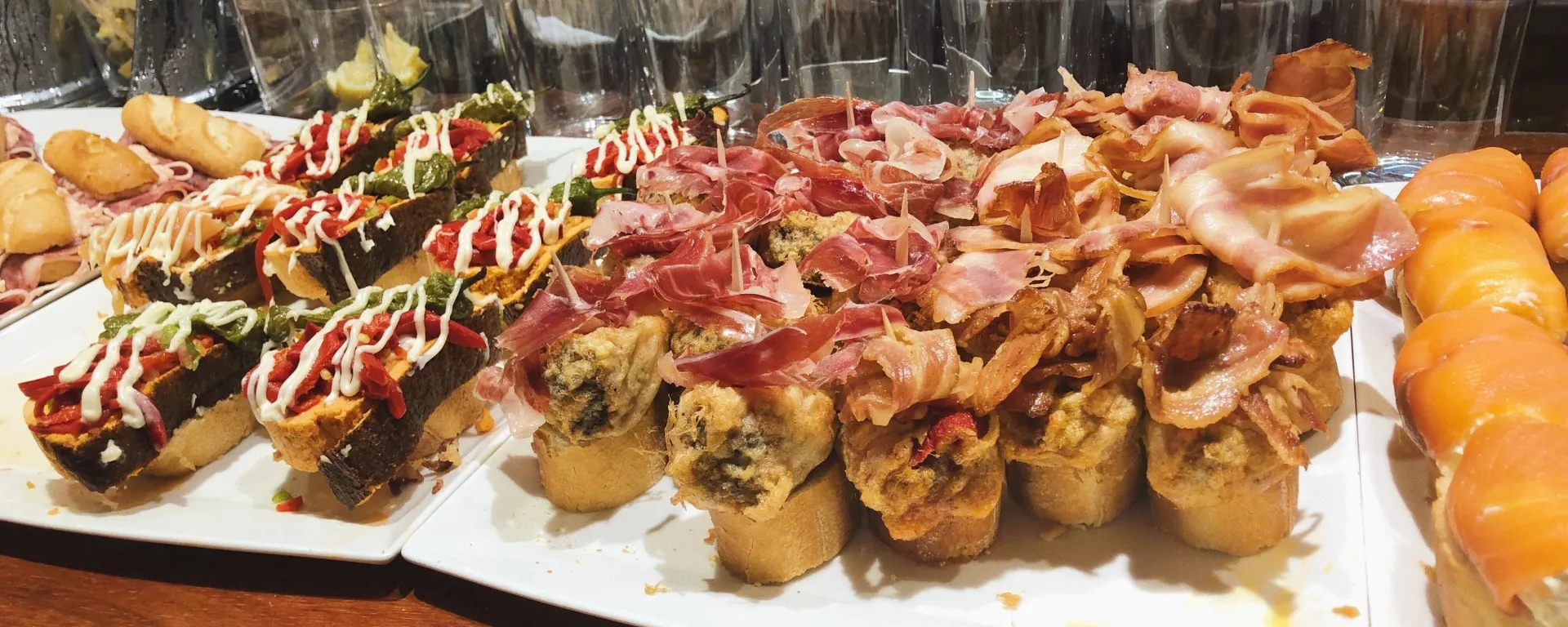 Pintxos crowded on white platters along a bar counter: toasts of red pepper and green guindilla with ribbons of mayonnaise, others under folds of cured ham and crisp bacon, and rolls of smoked salmon at the right, with glasses on the bar behind