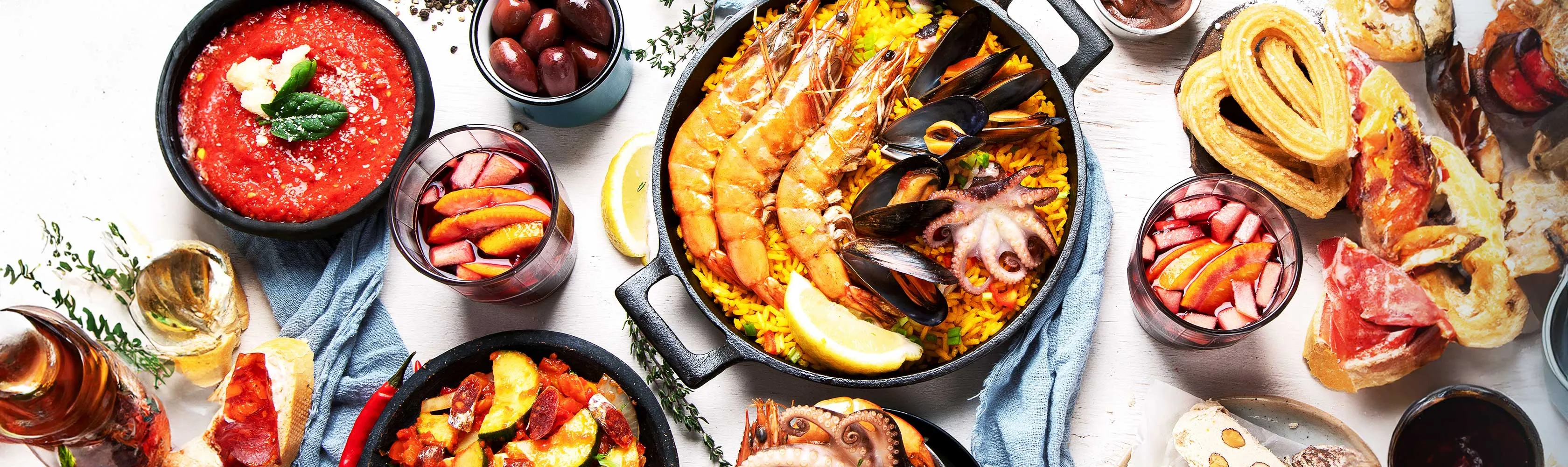 Spanish dishes from above on a white table: a seafood paella with prawns, mussels and octopus, a bowl of tomato soup, glasses of sangria, churros, olives and cured ham