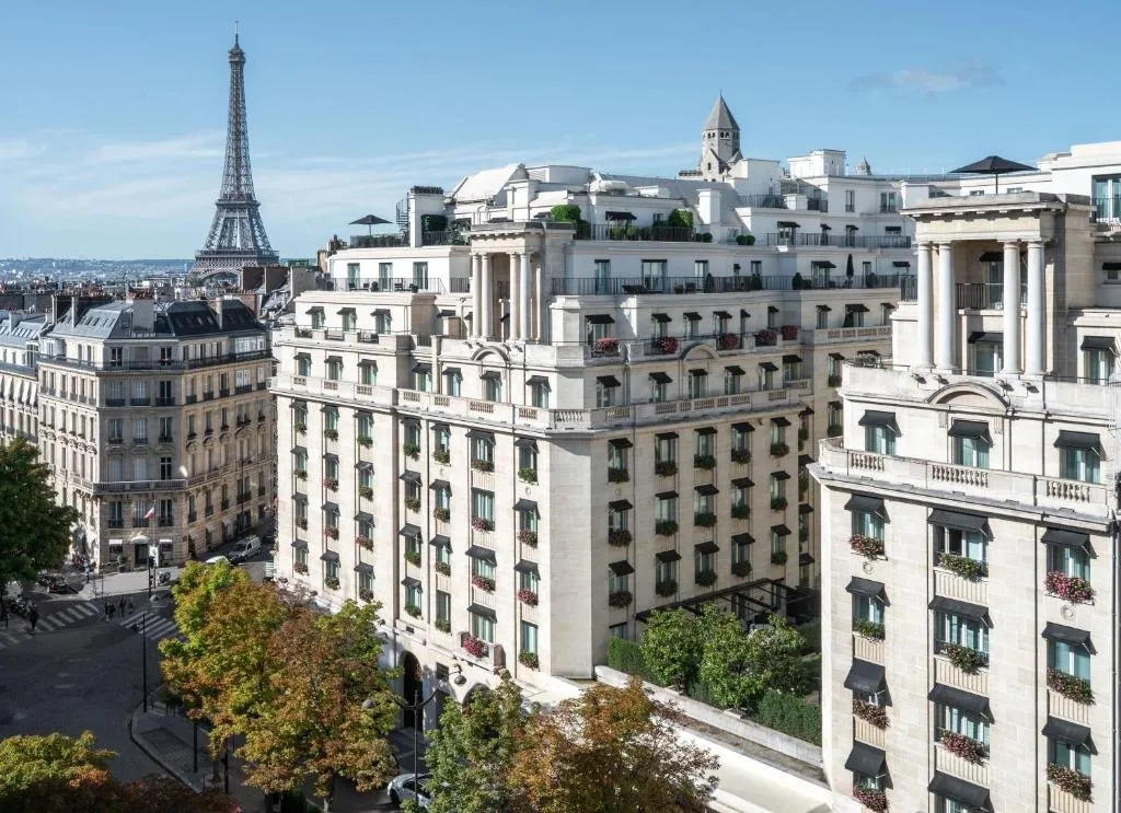 The hotel's pale stone building with red flowers in its window boxes and pavilions on the roof, above trees on a street corner, with the Eiffel Tower beyond