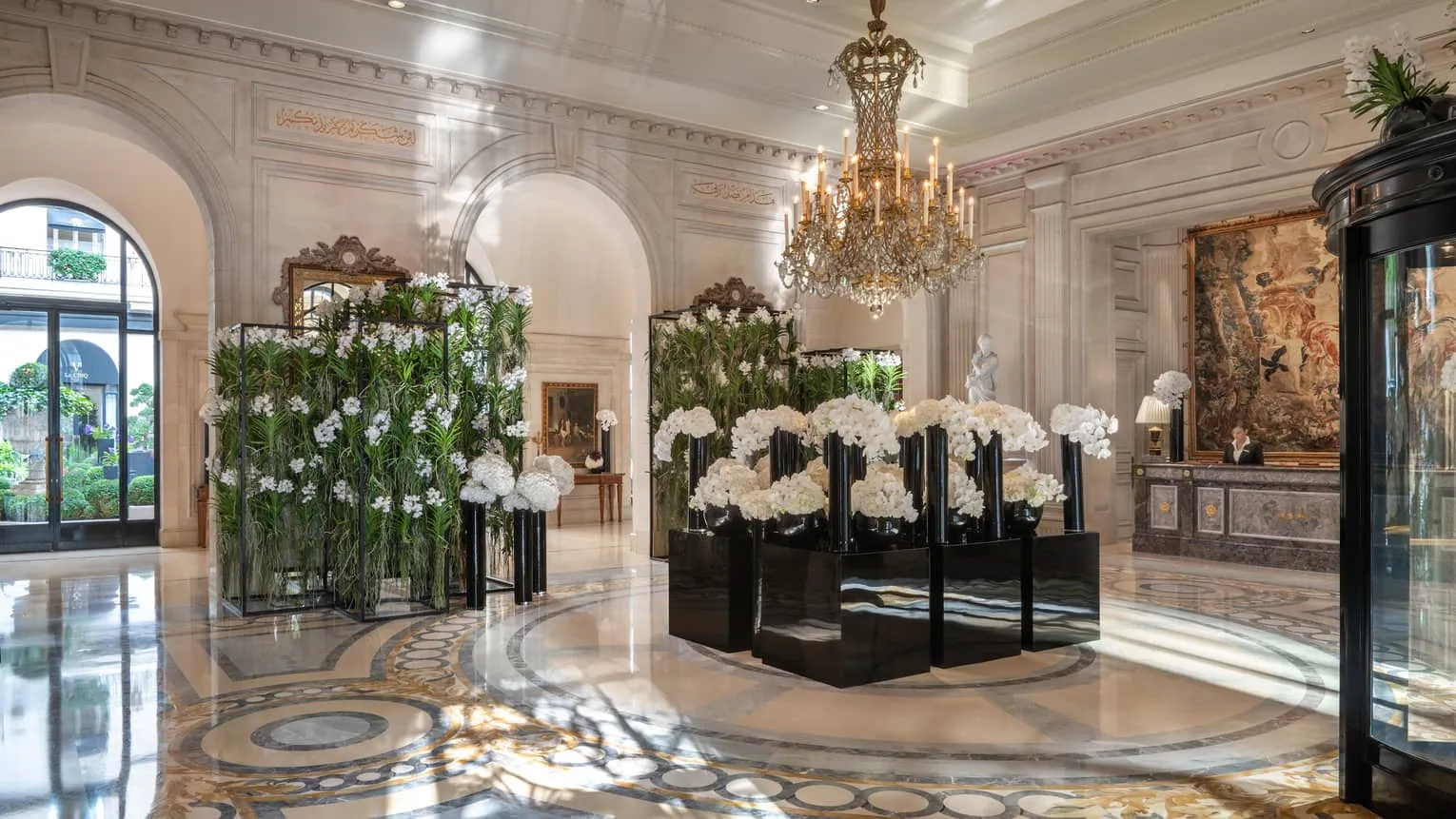 The hotel's marble lobby: a crystal chandelier with candle lights above black vases of white orchids and hydrangeas, glass cases full of green orchids, a tapestry behind the reception desk, and an arched glass door at the left with plants beyond it