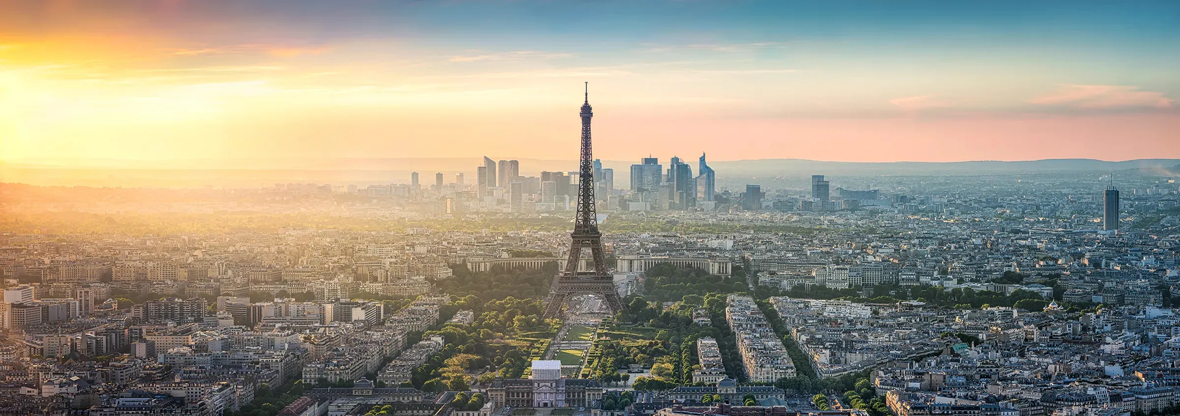 Paris from high above at sunset: the Eiffel Tower rising from the green Champ de Mars, the city's pale rooftops spreading on every side, and the towers of La Défense on the hazy horizon under a gold and blue sky