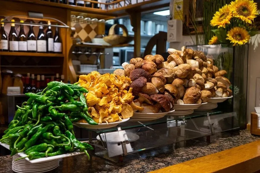 A bar counter heaped with wild mushrooms, golden chanterelles beside plates of ceps on a glass shelf, and a pile of green peppers, with wine bottles on the shelves behind and sunflowers in a vase
