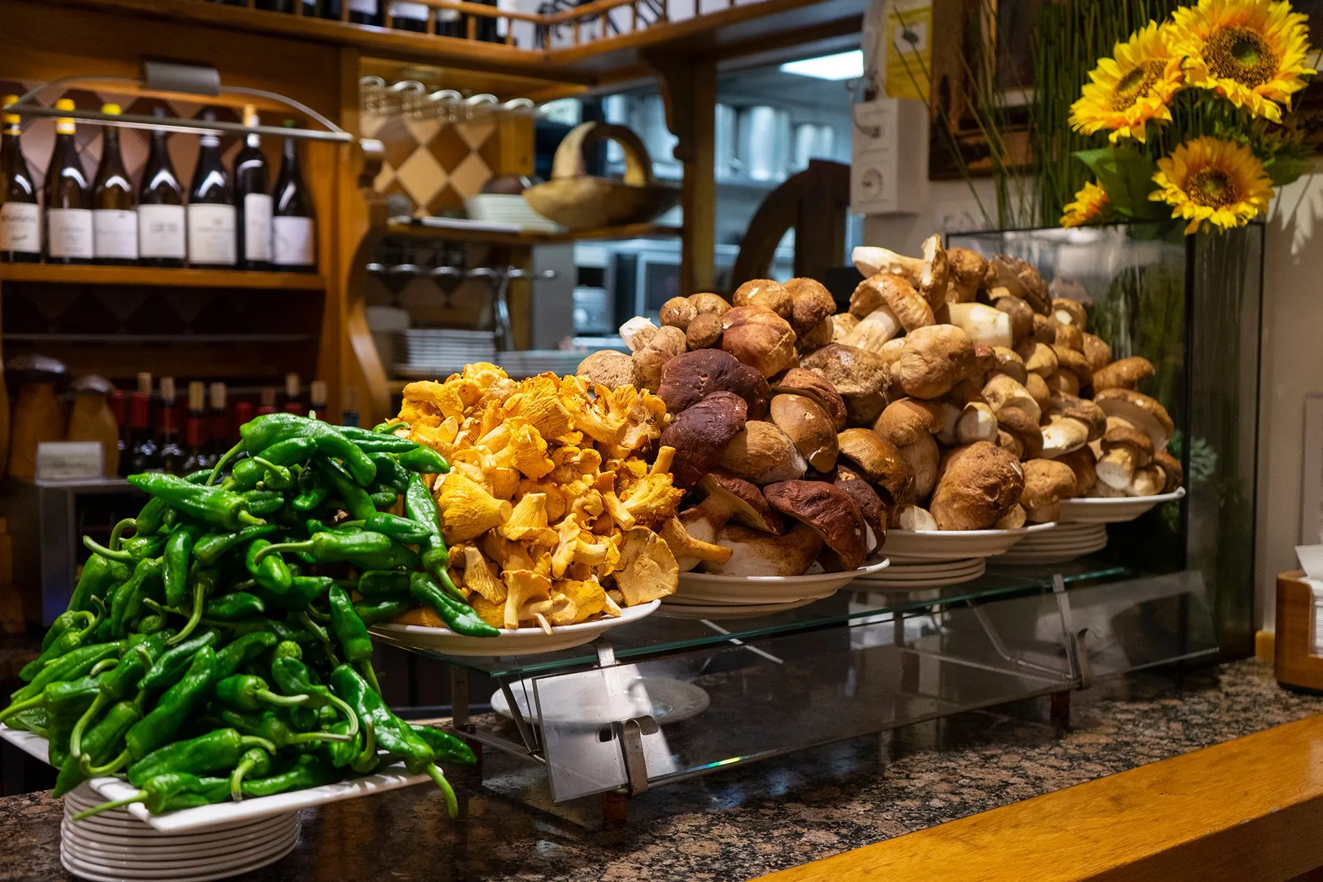 A pintxo bar counter heaped with wild mushrooms, golden chanterelles beside plates of ceps, and a pile of green peppers, with wine bottles on the shelves behind and sunflowers in a vase
