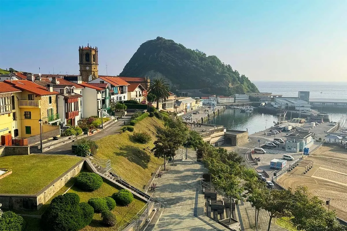 Getaria from above in low sun: red-roofed houses and a stone church tower on the left, gardens and steps running down to the harbour, and a wooded headland rising beyond the moored boats, with the sea to the right