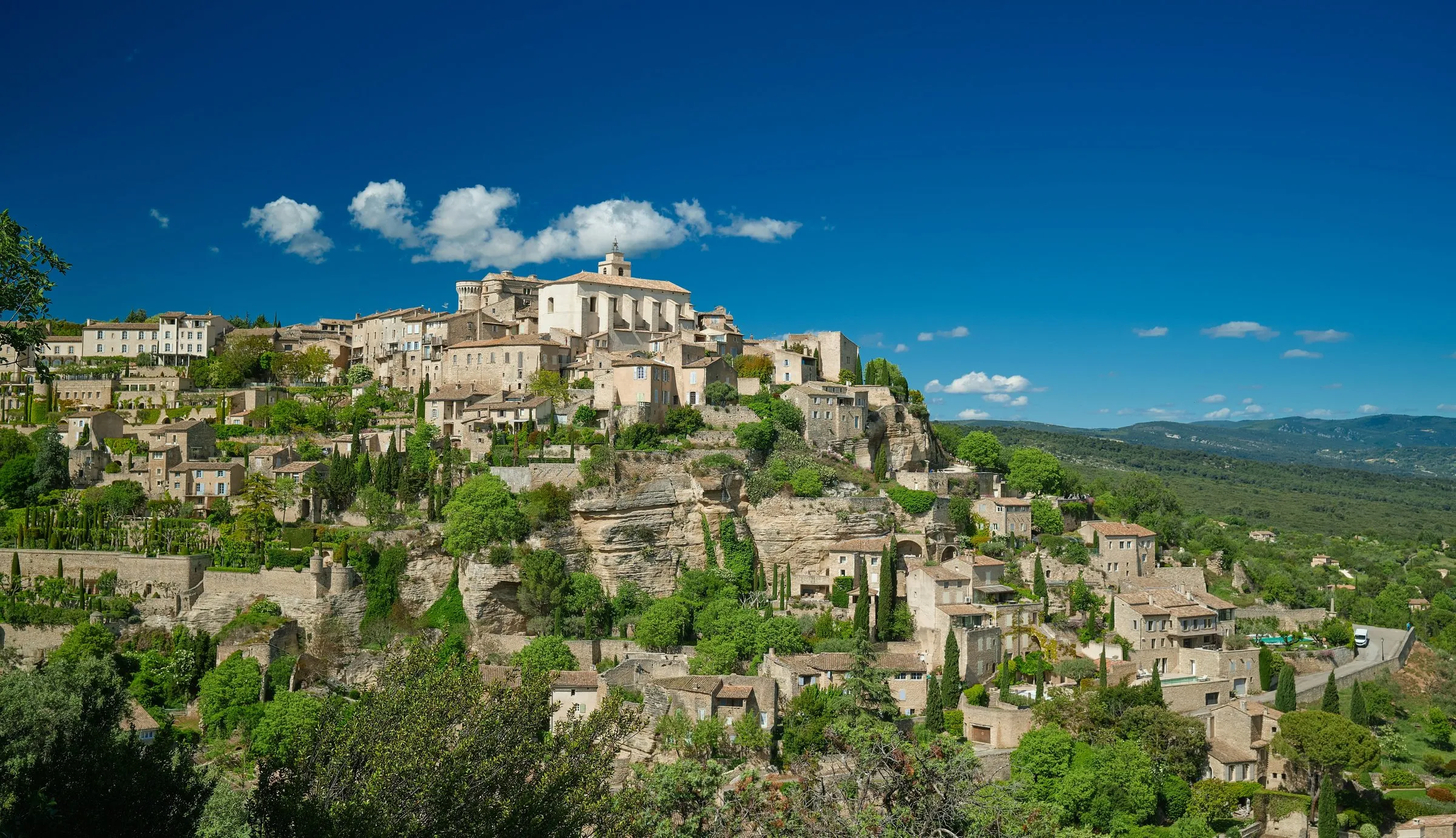 Gordes from the belvedere: the château and the church of Saint-Firmin on the summit, stone houses stacked down the cliff among cypresses, the Luberon foothills beyond under a deep blue sky with small clouds