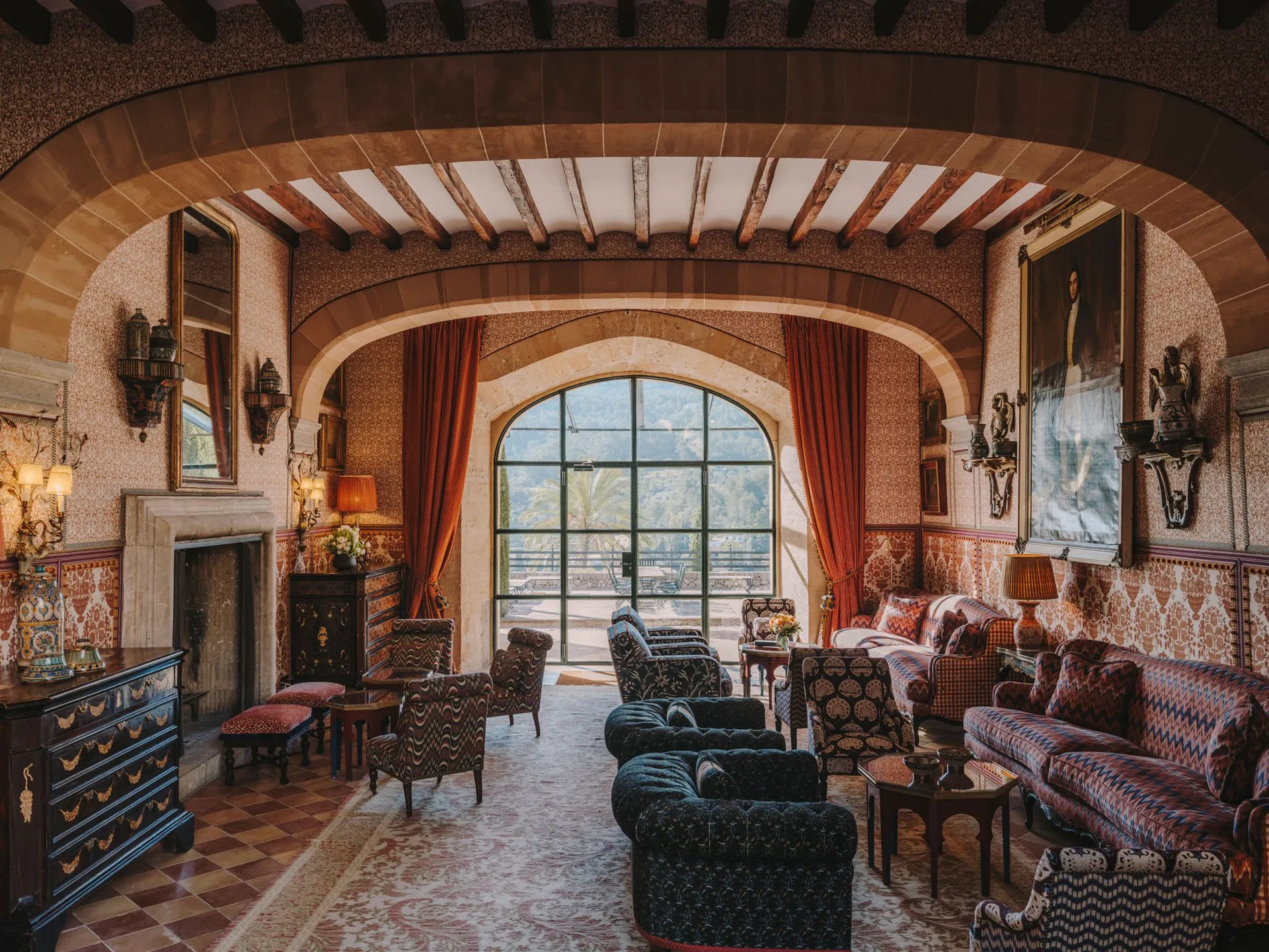 Sitting room at Grand Hotel Son Net under a stone arch and beamed ceiling, with a fireplace and an arched glass door onto a terrace and wooded hills