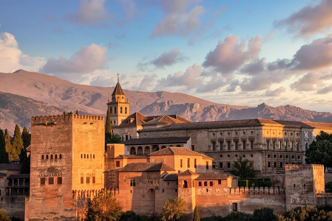 The Alhambra's walls lit gold in the late afternoon: a tall crenellated tower on the left, a church bell tower and the Palace of Charles V with its round windows behind, and the bare ridges of the Sierra Nevada beyond under scattered cloud