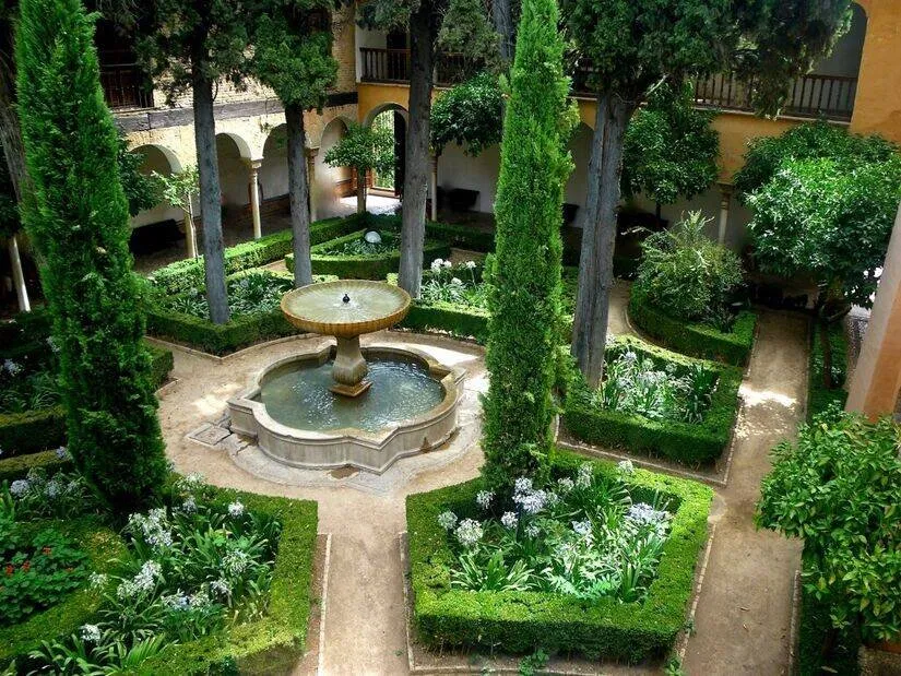A walled garden courtyard in the Alhambra seen from above: a fountain with a raised bowl in a lobed basin, beds of white flowers edged with clipped box, tall cypresses, and an arcaded gallery behind