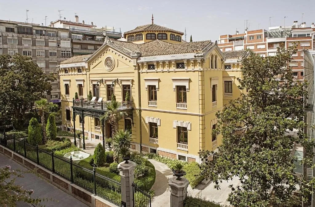The palace from above on a clear day: a yellow villa with a pediment and a round window, a cast-iron porch carrying the main balcony and an octagonal lantern on the tiled roof, behind iron railings and a garden of palms, clipped cypresses and a small fountain, with taller apartment blocks around it