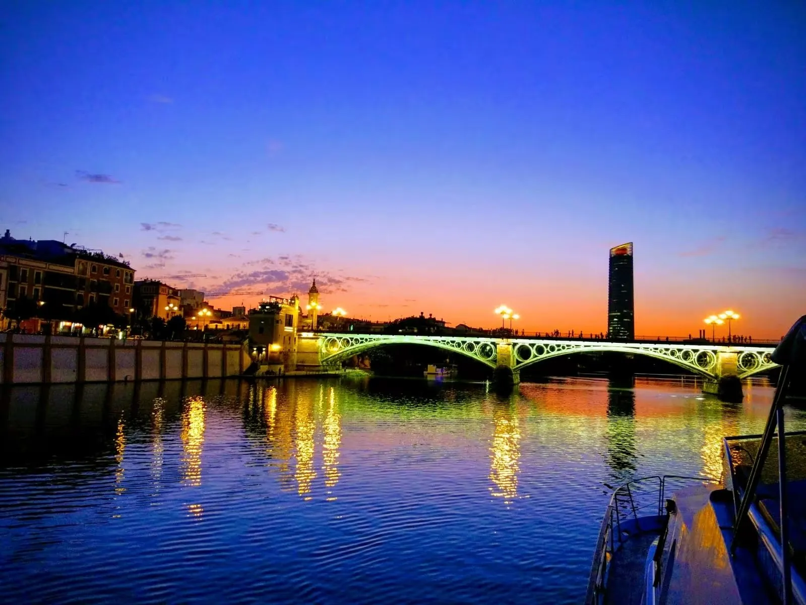 The Triana bridge lit at dusk over the Guadalquivir, the Torre Sevilla beyond and a boat's bow in the foreground