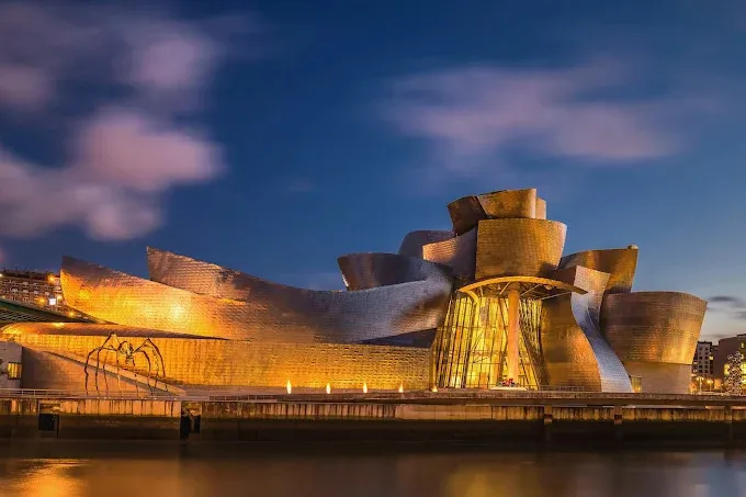 The Guggenheim Museum Bilbao lit gold at dusk, its curving metal-clad forms above the river, with a giant spider sculpture at the left