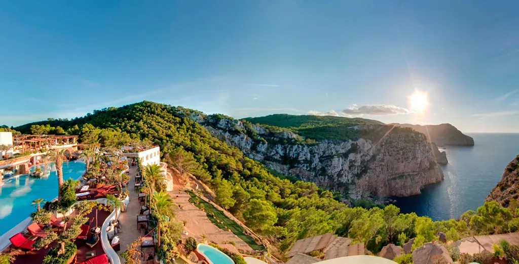 A white hotel and its terraces of pools, palms and loungers on a pine-covered cliff top, the cliffs of a wooded headland falling to a deep blue sea in low evening sun