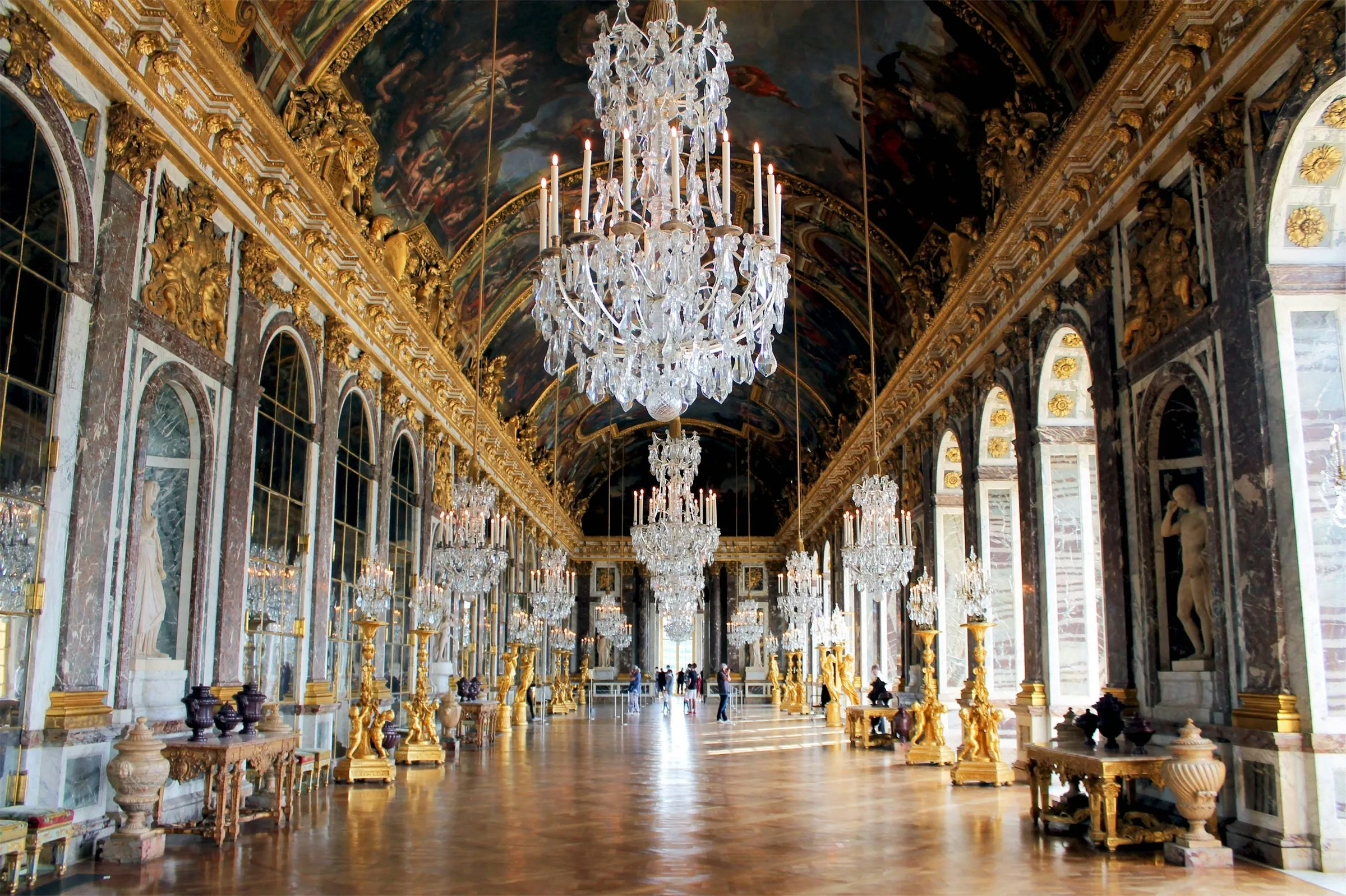 The Hall of Mirrors at Versailles seen down its length: crystal chandeliers hanging from the painted vault, gilded candelabra and marble statues along both sides, and a few visitors far down the parquet