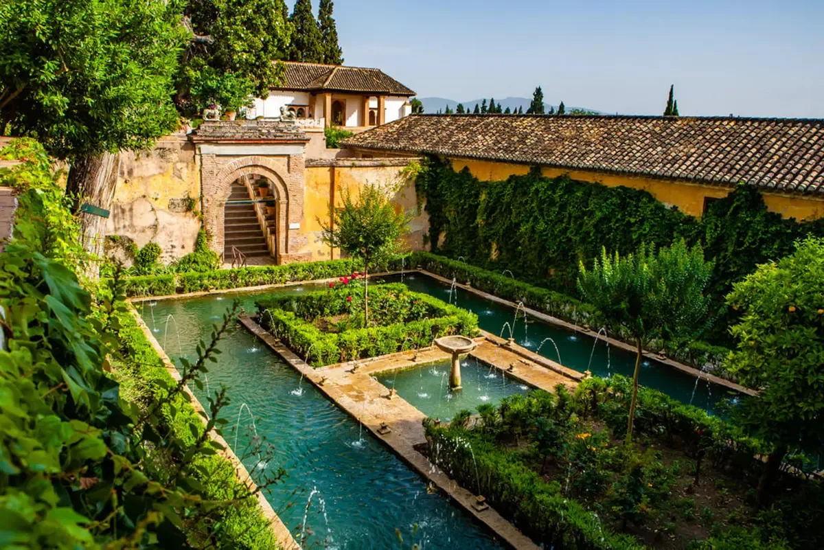 A garden court with a long pool lined with fountain jets around a central island of clipped hedges, white walls with an arched doorway and steps behind, under tall cypresses