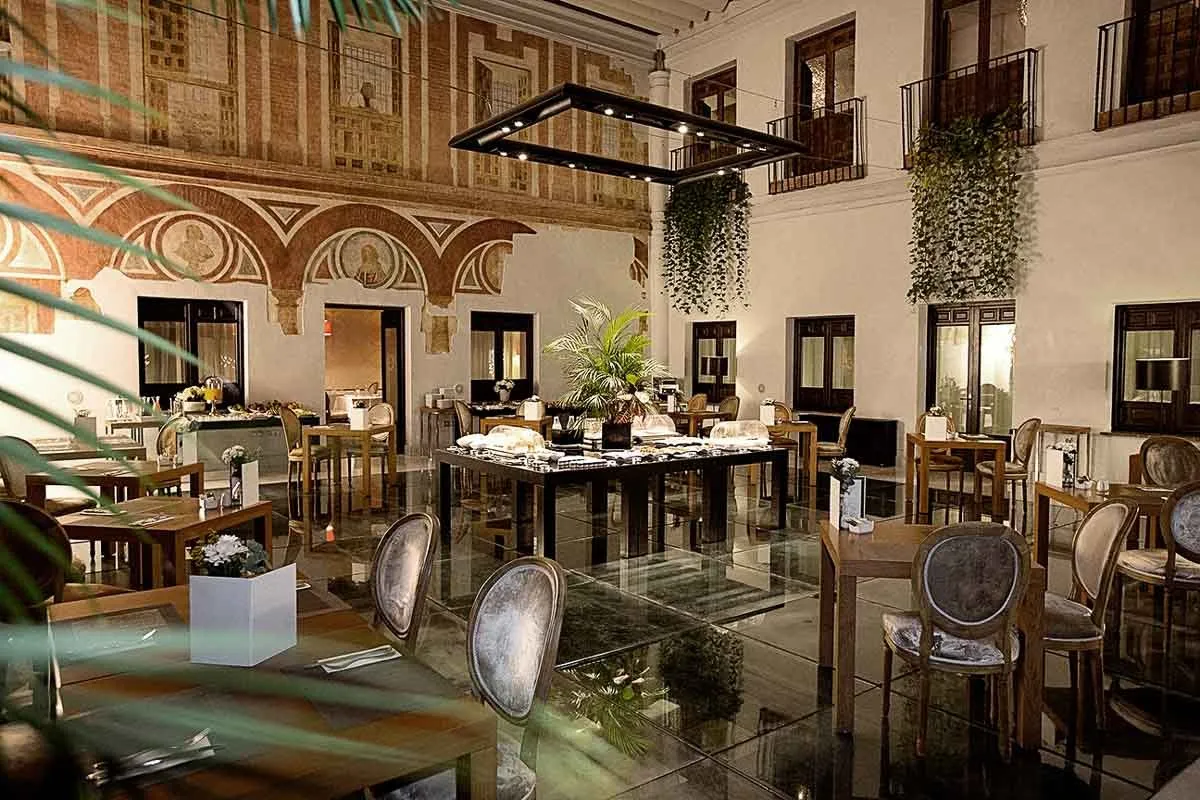 A courtyard at night with its tables laid: grey velvet chairs with oval backs on a glass floor, a long buffet table at the centre under a square frame of small lights, ivy trailing from iron balconies, and a wall painted with red and ochre arches and portrait roundels