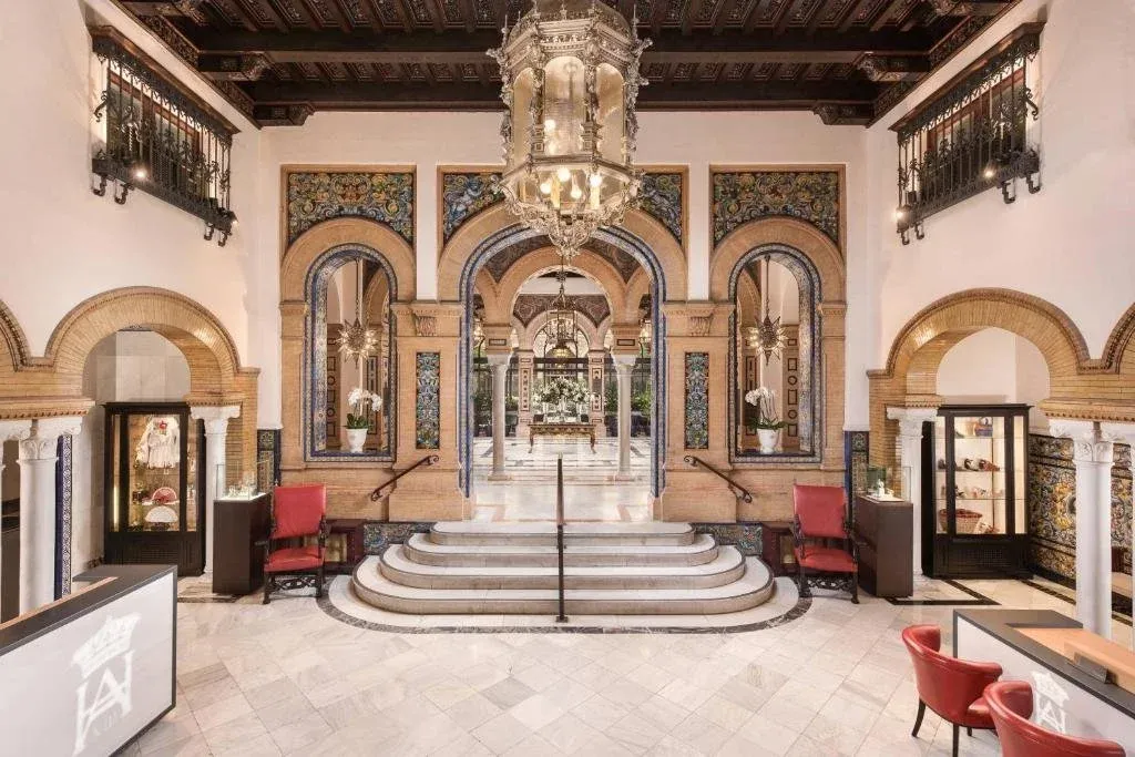 The entrance hall of Hotel Alfonso XIII: brick horseshoe arches with painted tilework, a large glass lantern hanging from a coffered wooden ceiling, and marble steps leading through to the courtyard beyond
