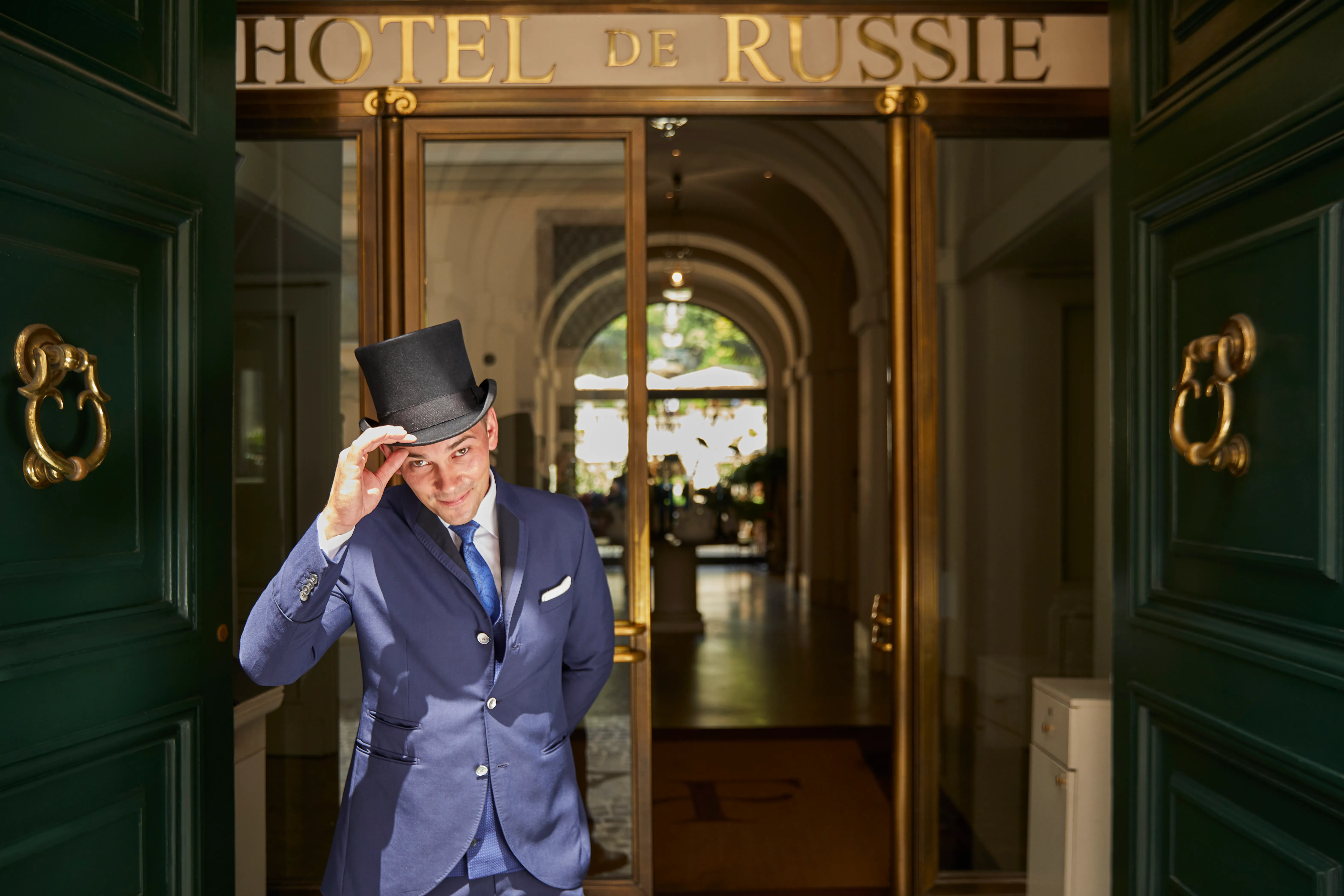 The doors of Hotel de Russie on Via del Babuino, a doorman in a top hat between them and the garden seen through the hall beyond