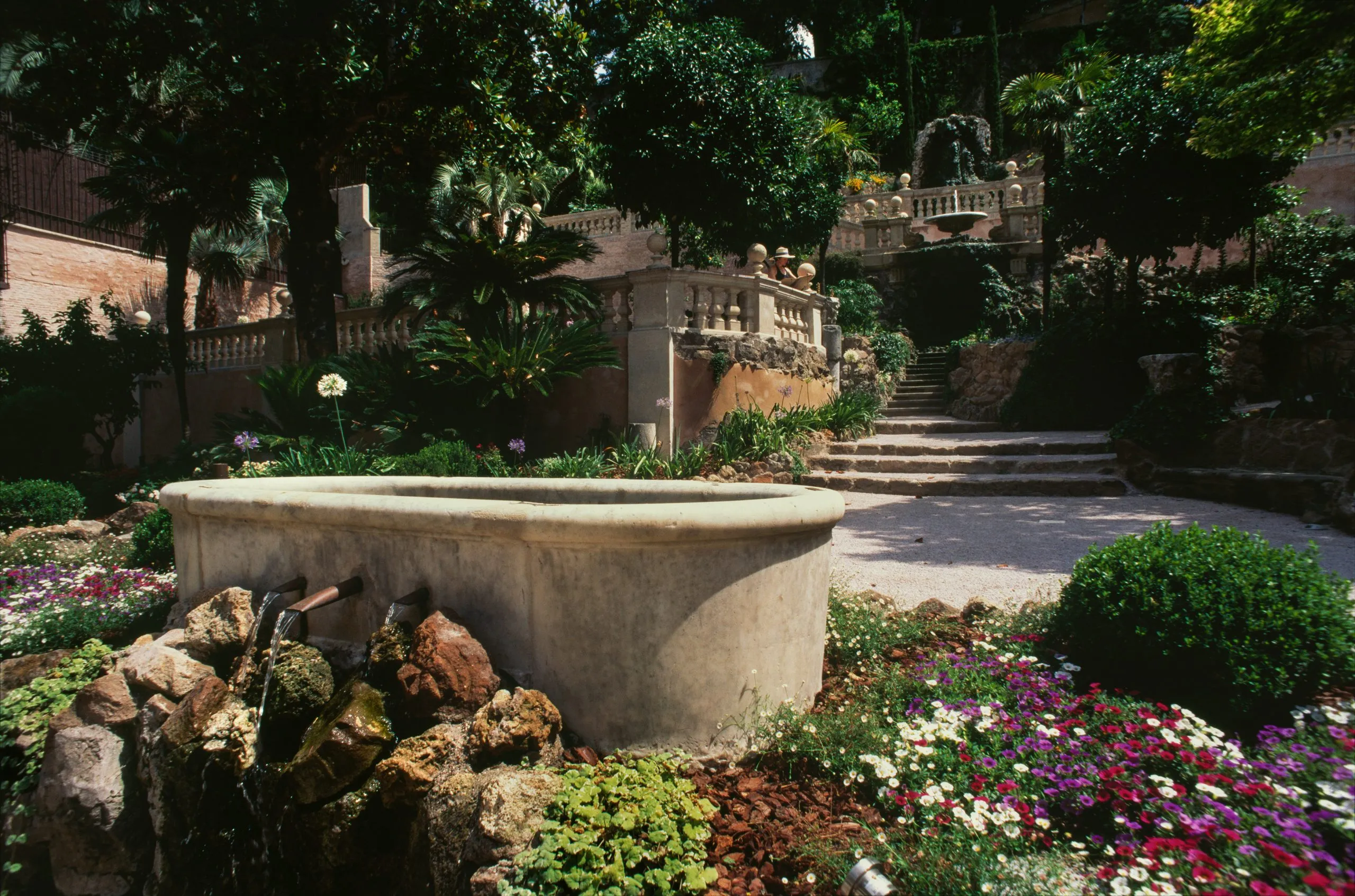 A terraced garden in full sun: an oval stone basin fed by two spouts among rocks and flowers in the foreground, box and agapanthus around it, a balustraded stair climbing between palms, pines and orange trees to a fountain grotto above
