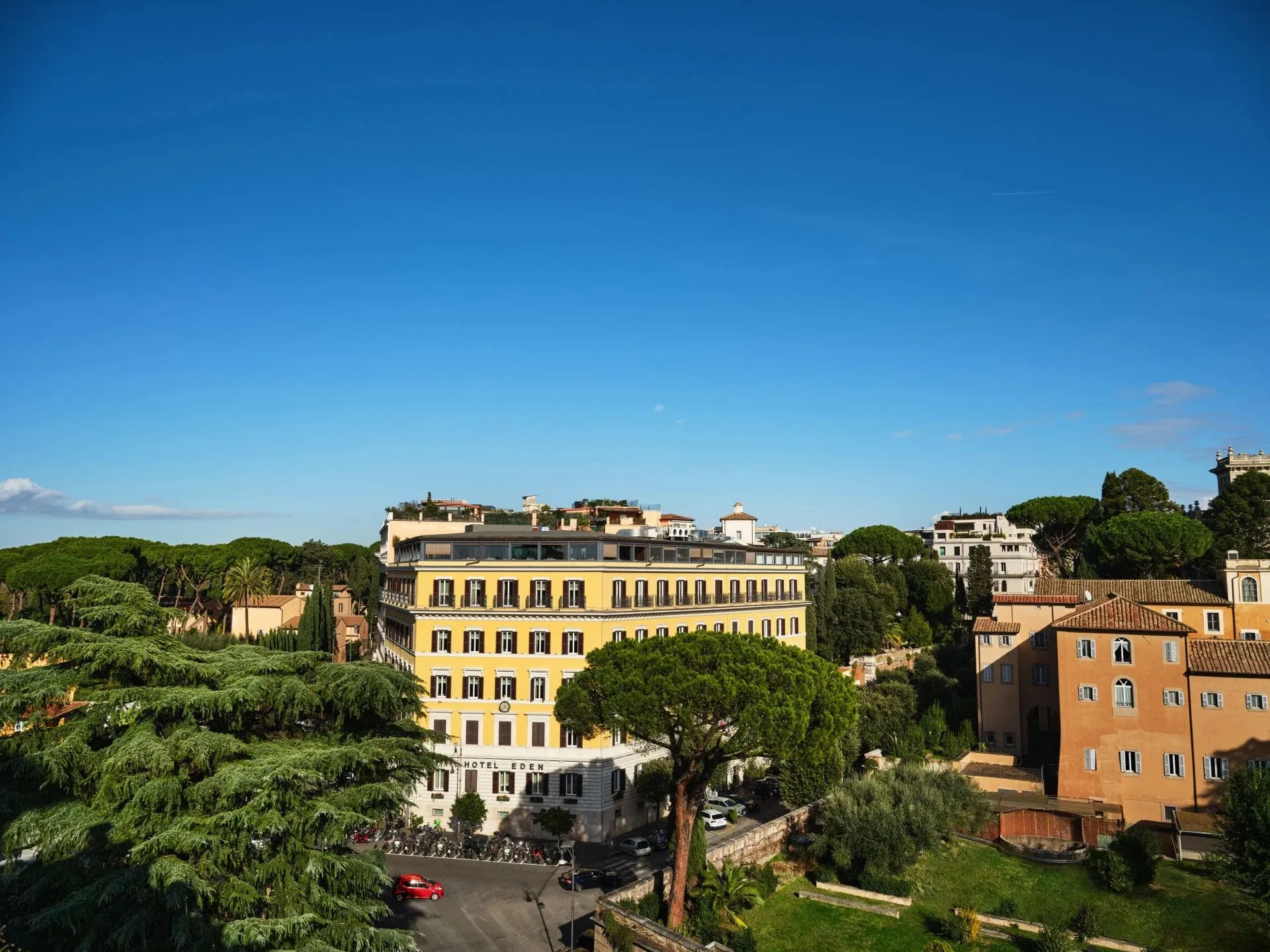 Hotel Eden from across the street on the Ludovisi hill: the yellow hotel with its glazed rooftop floor among pines and cedars, ochre villas on the hill behind, a clear blue sky