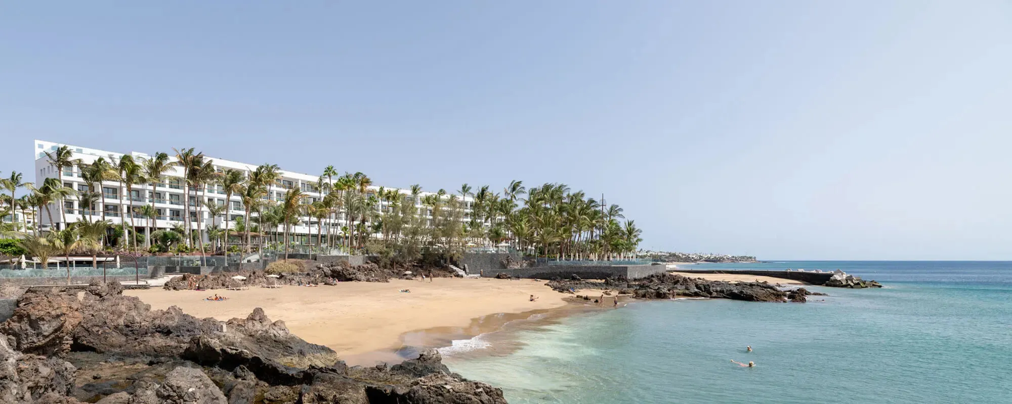 Hotel Fariones, a low white building behind tall palms, above a sandy cove edged with black volcanic rock and clear turquoise water