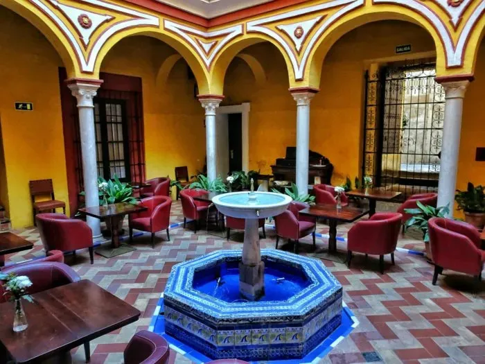 A covered patio at Las Casas de la Judería: yellow and red scalloped arches on marble columns around a blue-tiled octagonal fountain, with tables and red tub chairs set out on herringbone brick