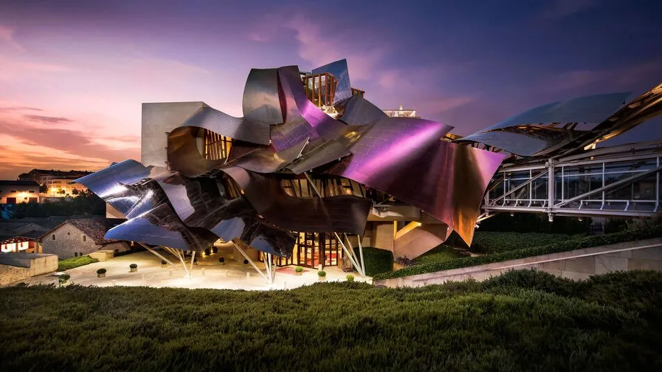 The Hotel Marqués de Riscal at dusk: ribbons of titanium in purple, pink and silver curling over the building on slender pillars, a glass footbridge to its right and a lawn in the foreground