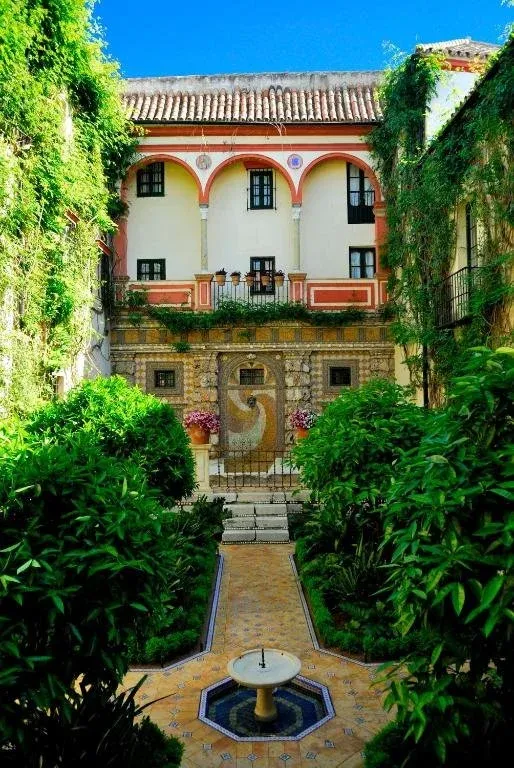 A planted patio at Las Casas de la Judería, its arcaded upper gallery above a tiled wall fountain, with clipped shrubs and a small stone fountain in the tiled floor