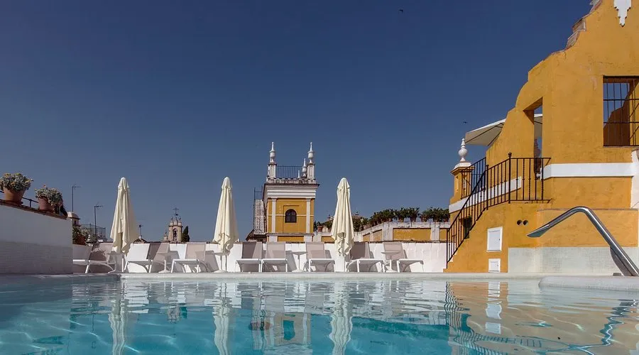 The rooftop pool in full sun, loungers and folded parasols along its far side, with a small yellow belfry and the rooftops of the quarter beyond