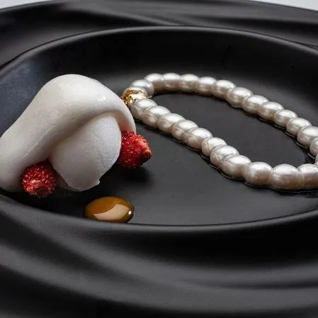 A white, folded shape on a black plate with two wild strawberries, a drop of amber sauce and a ring of pearls finished with a gold clasp