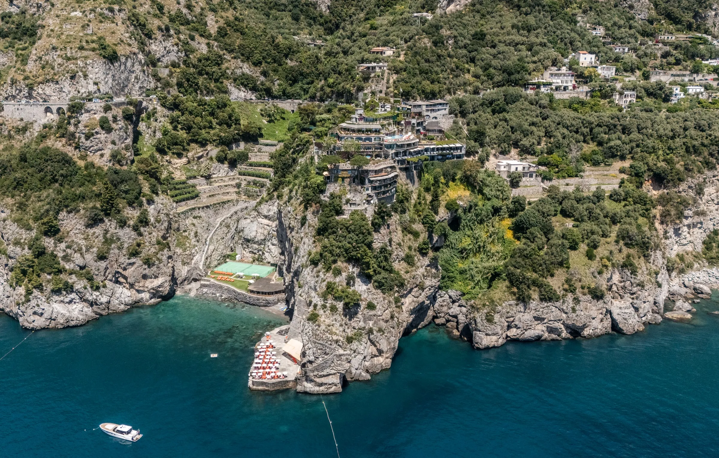 The whole headland from the air over the sea: the hotel's terraces and pool at the top of the promontory, the green tennis court on a shelf halfway down, terraced gardens, the coast road's arched viaduct top left, the beach club at the foot of the rock with ro