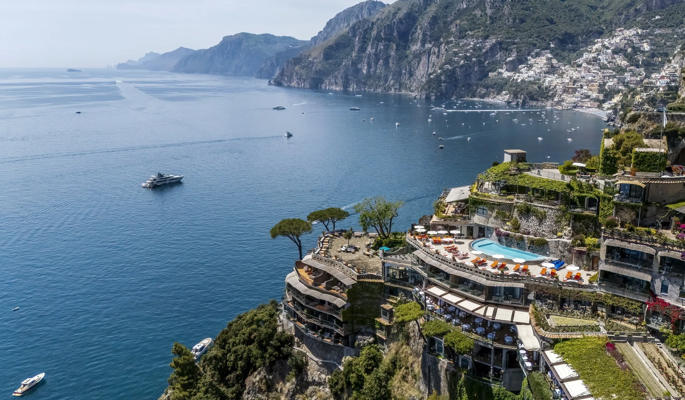 The hotel from the air above the sea: its terraces stepped down the tip of the wooded promontory, the pool with orange loungers on the upper terrace, a stone pine, dining tables below; the coast runs away to the left, Positano's houses up the hillside top righ
