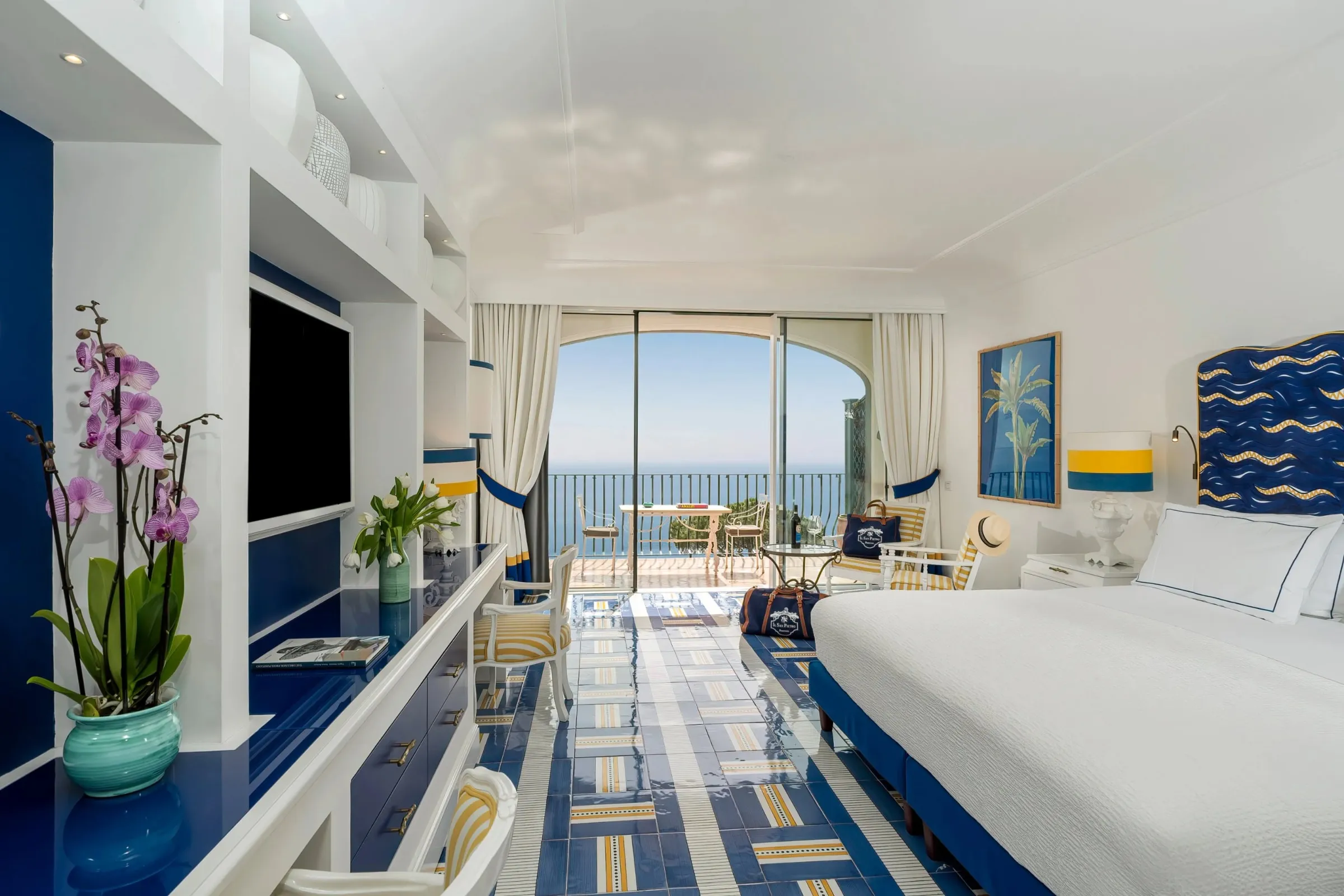 A Premier room from the entrance: white vault, blue-lacquered cabinets and shelves with a television, an orchid, blue-and-yellow majolica floor, white bed with a blue wave headboard; through the open glass door a terrace with a white table and chairs at a rail