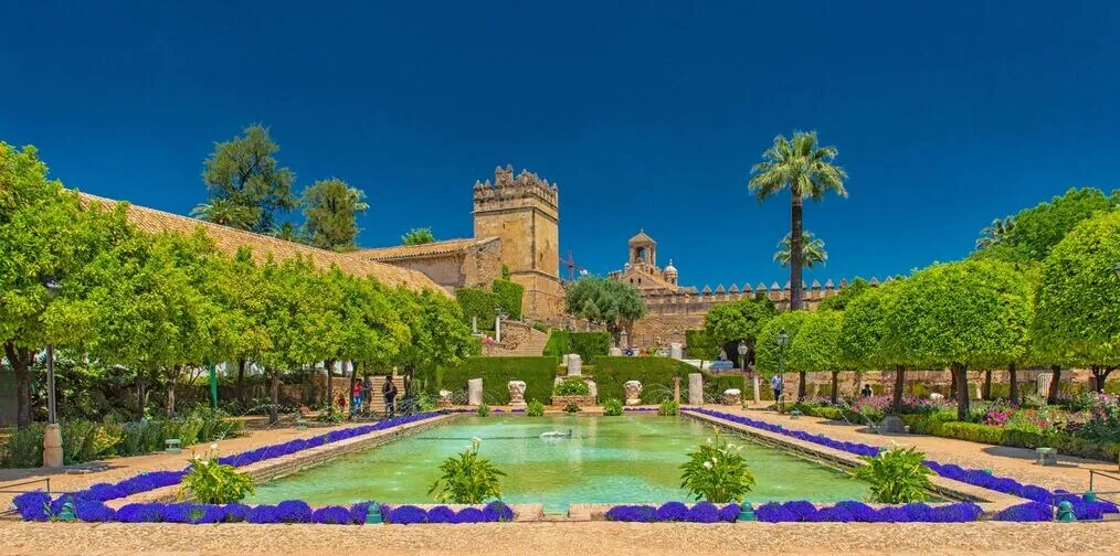 The gardens of the Alcázar de los Reyes Cristianos: a long pool edged with violet flowers between clipped trees and palms, and the fortress's crenellated stone tower behind it under a deep blue sky