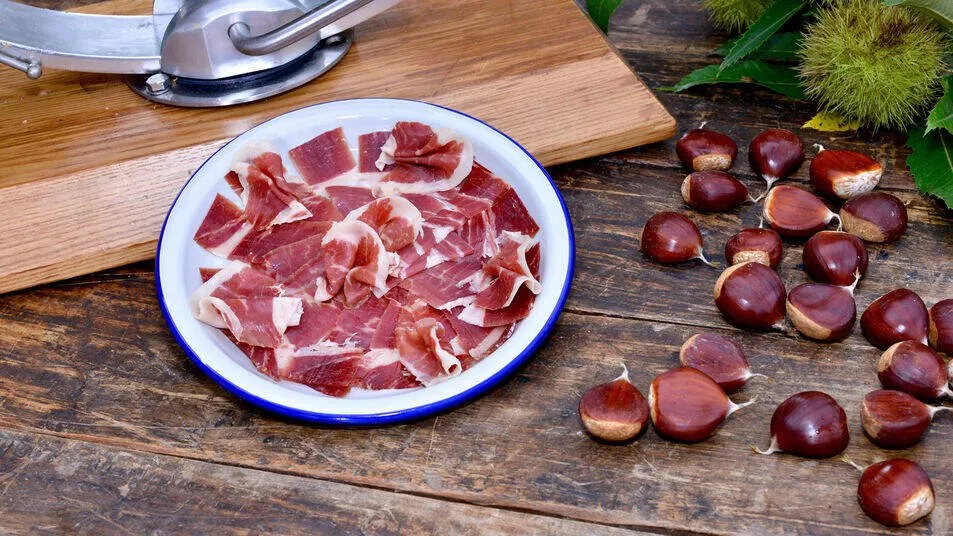 Thin slices of jamón ibérico on an enamel plate on a wooden table, with a ham stand behind and chestnuts scattered beside it